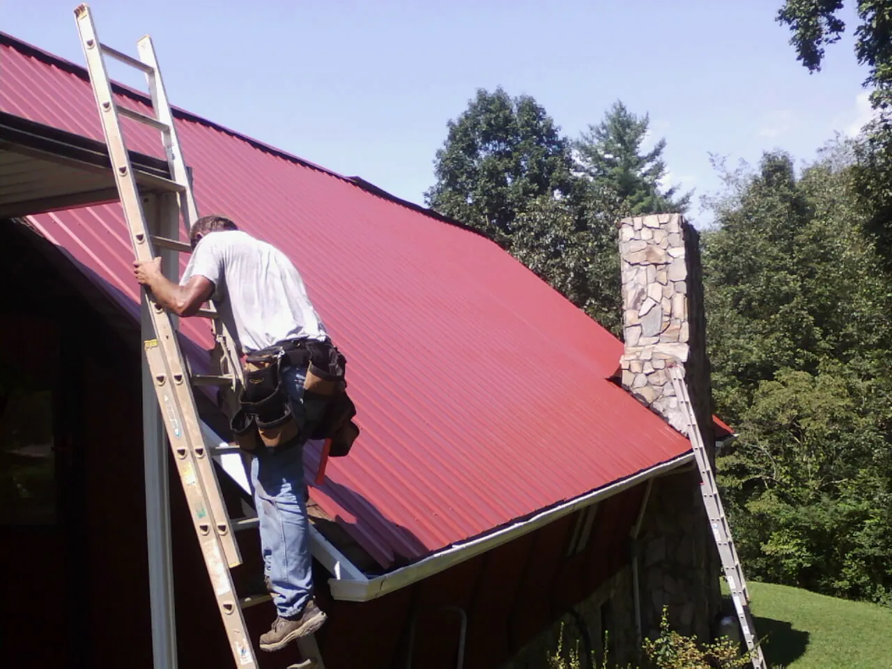 Roofer on a ladder beside a red metal roof and stone chimney — Soffit & Fascia Repair by Roofing Brothers in Grants Pass